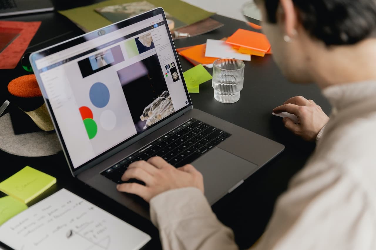 An individual works on a MacBook displaying a colorful graphic design project, with an organized desk featuring sticky notes, a notebook, a glass of water, and a decorative plant.