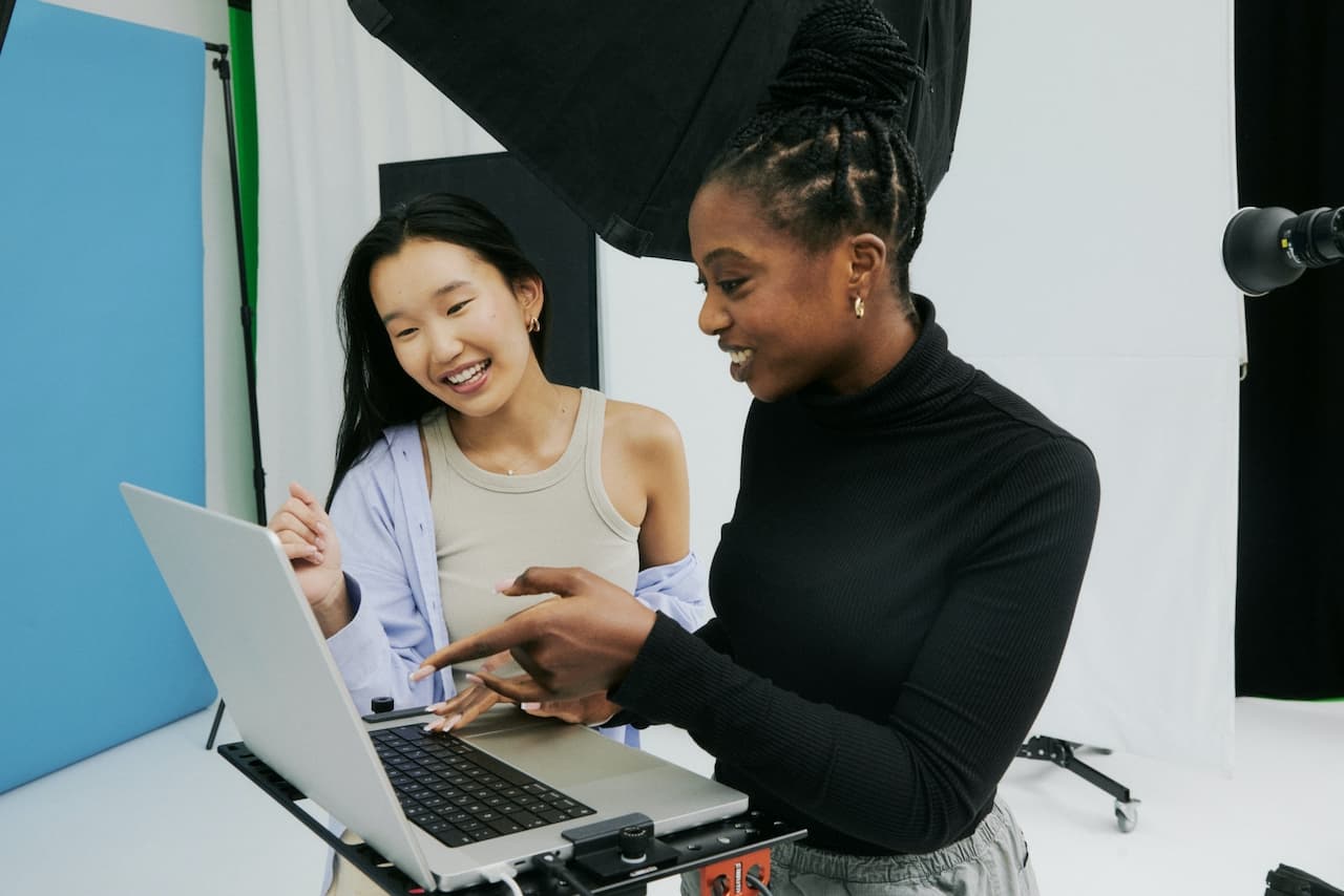 Two smiling people collaborating on a laptop in a photography studio with lighting equipment and a blue backdrop visible.